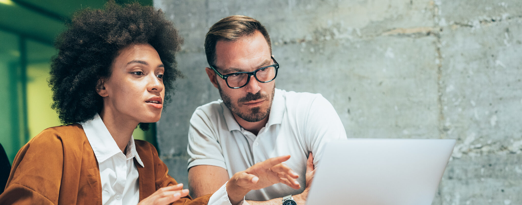 Two colleagues in an office are sitting at a desk and talking to each other while looking at a laptop.